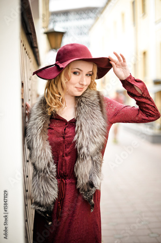 Красивая девушка со светлыми волосами стоит на улице в бордовом платье и шляпе с меховым воротником  © natasidorova 