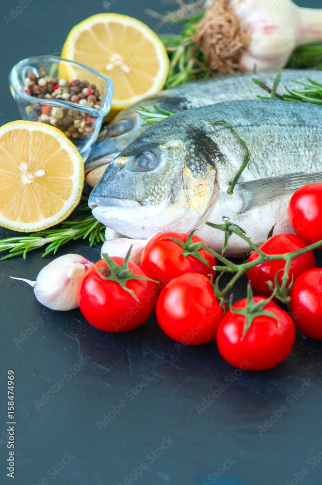 Fresh raw dorado fish with rosemary, garlic, tomatoes, pepper and lemon on a black slate background. Selective focus. Close up. Fresh raw dorado fish with rosemary, garlic, tomatoes, pepper and lemon on a black slate background. Selective focus. Close up.