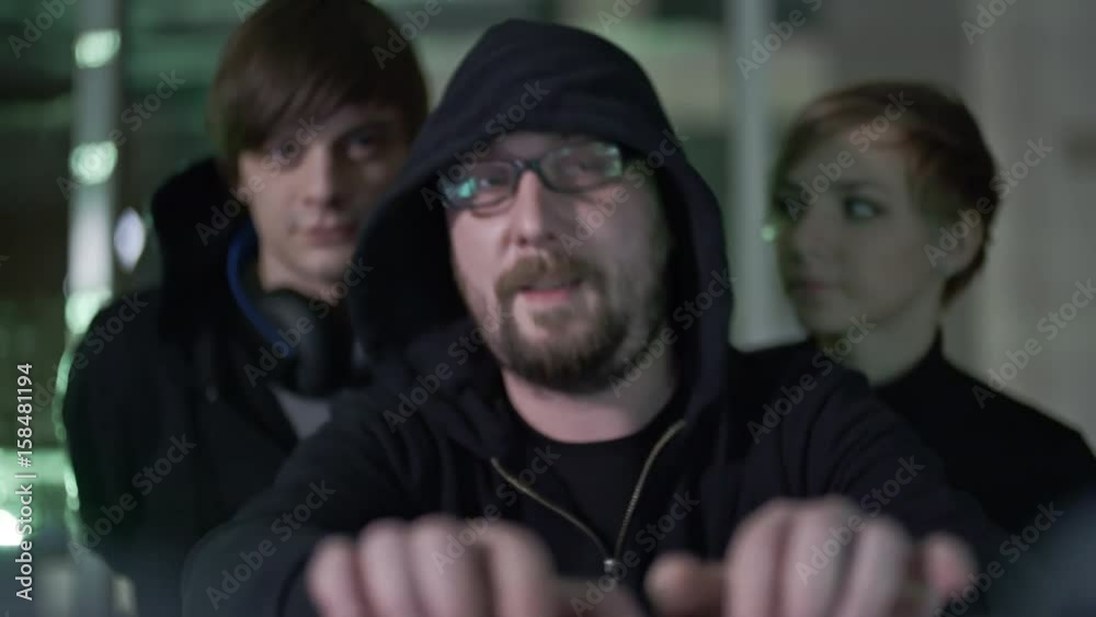 Bearded hacker in eyeglasses and hood working on computer with two teammates standing behind his back and discussing something in dark room Bearded hacker in eyeglasses and hood working on computer with two teammates standing behind his back and discussing something in dark room