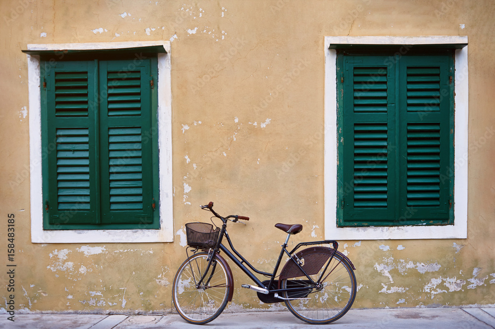 Bicycle with metal basket parked in front of an old wall of a house with flaked yellow plaster and big widows with green shutters