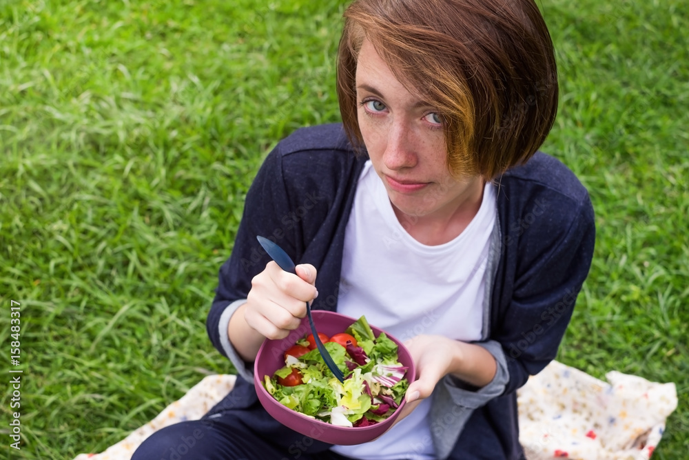 A girl sits on the grass and eats a green fresh salad with plastic folk. A healthy diet on plant foods. A girl sits on the grass and eats a green fresh salad with plastic folk. A healthy diet on plant foods.