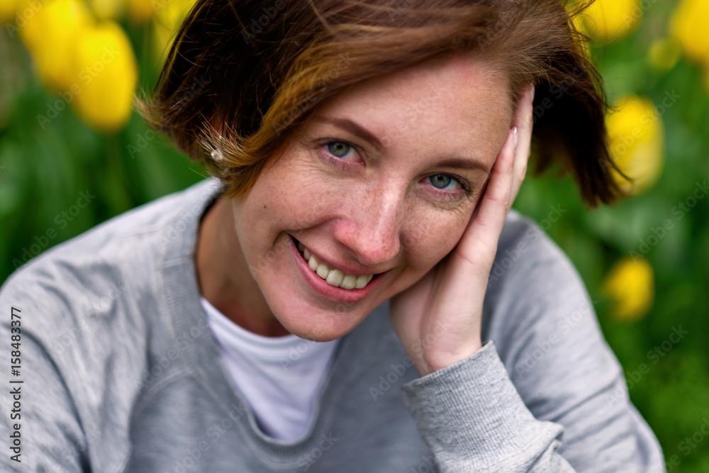 A girl with freckles sits on the grass beside the yellow tulips in the park.