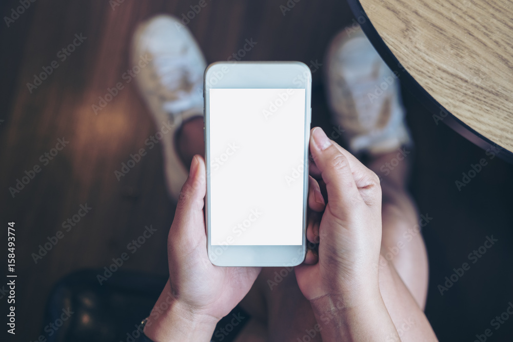 Mockup image of woman's hand holding white mobile phone with blank screen on thigh with wooden floor background in modern cafe