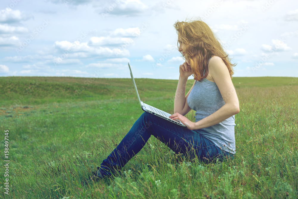Woman sitting on a green meadow on the background of sky with clouds and working or studying with laptop wireless Woman sitting on a green meadow on the background of sky with clouds and working or studying with laptop wireless