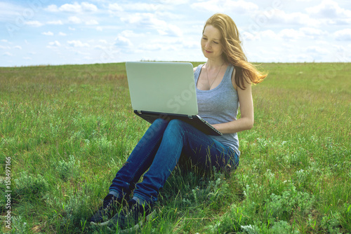 Woman sitting on a green meadow on the background of sky with clouds and working or studying with laptop wireless