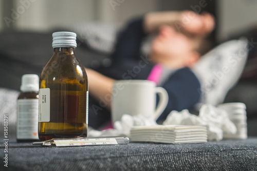 Sick woman holding head and forehead with hand and arm. Medicine, thermometer, hot beverage and dirty paper towels in front. Person having flu, fever and headache on sofa couch. Caught cold in winter. photo