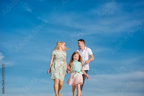 Happy beautiful laughing family running along the sand on the beach against the background blue sky in summer vacation