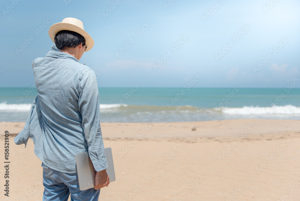 Young Asian happy man holding laptop on the beach, working outdoor in summer season, digital nomad lifestyle concepts