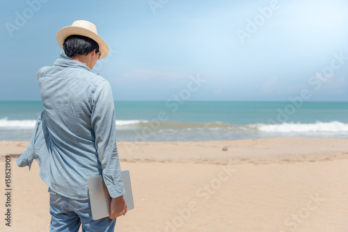 Young Asian happy man holding laptop on the beach, working outdoor in summer season, digital nomad lifestyle concepts