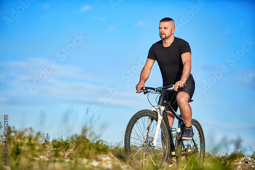 Young man cycling on a green meadow against blue sky with clouds.