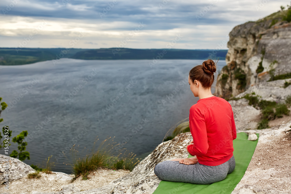 Rear view of young woman sitting in lotus position on the rock over the river. Rear view of young woman sitting in lotus position on the rock over the river.