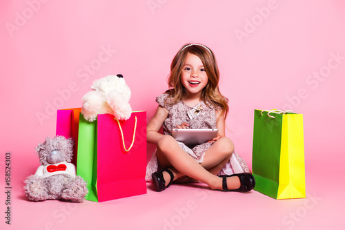 Little girl in a dress on a pink background with a teddy bear, shopping bags, and tablet