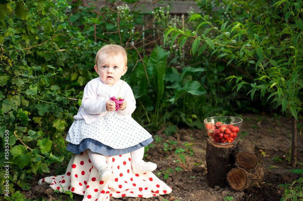 Girl 8 months old European Ukrainian little baby on a walk in the garden holds a flower and strawberries in her hands