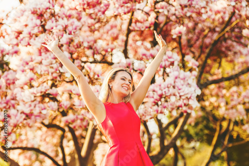 Beautiful happy young woman enjoying warm sunny day in spring garden  wearing bright red dress  arms wide open