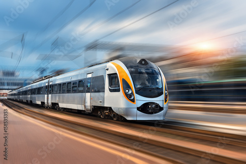 High speed train in motion at the railway station at sunset in Europe. Modern intercity train on the railway platform with motion blur effect. Industrial landscape with passenger train on railroad