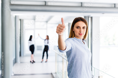Blonde business woman standing and showing thumbs up while there are her work colleague behind