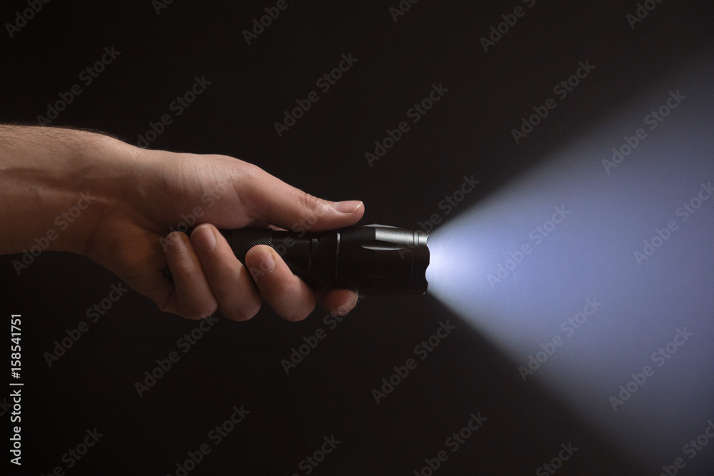 Black flashlight with wide beam in male's hand isolated from left side of the frame on black background - obrazy, fototapety, plakaty 