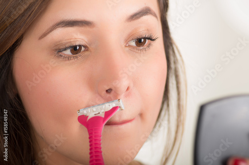 Close up of a beautiful young woman using a shaver pretending to shave her mustache looking at small mirror, in a bath background