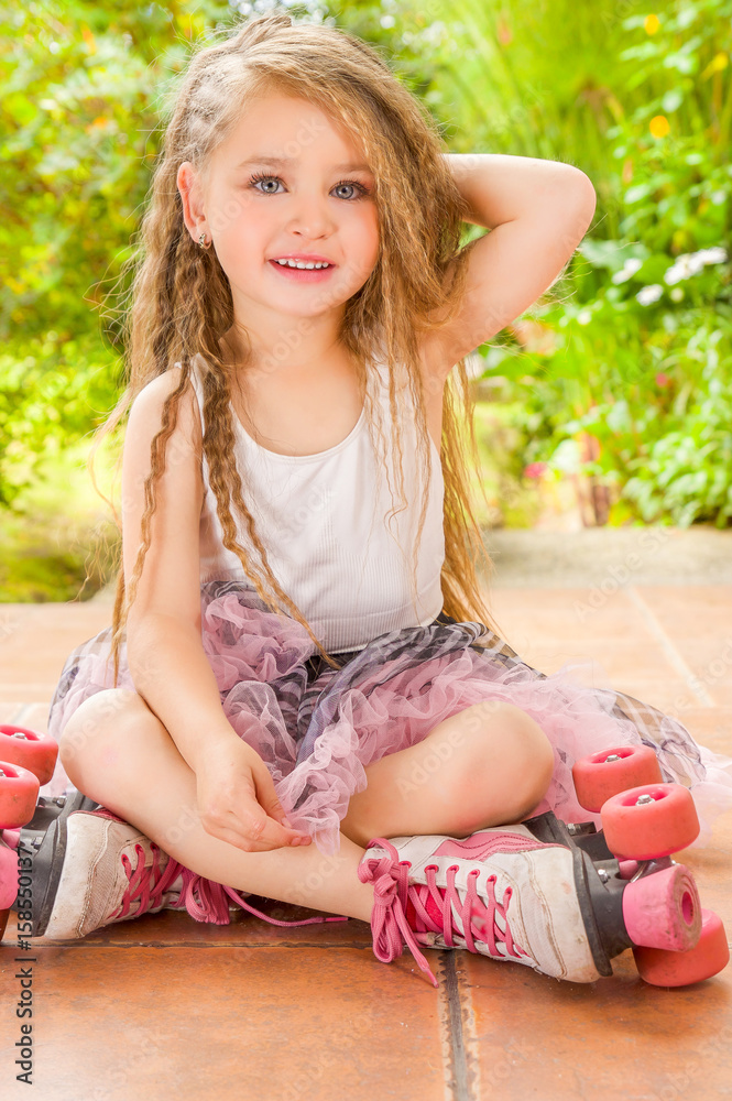 Little girl preschool sitting on ground wearing her roller skates and crossing her legs touching her head with one hand, in a garden background Stock-foto | Adobe Stock