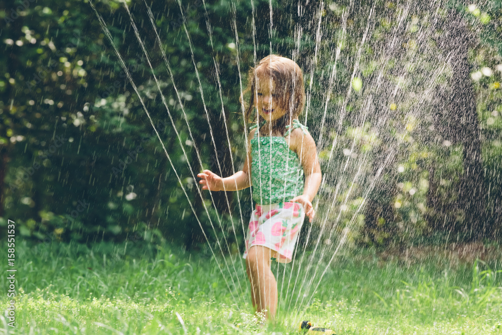 Happy toddler girl playing in a sprinkler - obrazy, fototapety, plakaty 