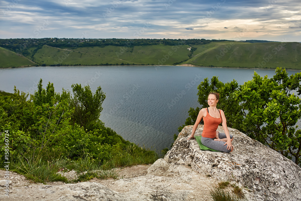 Beautiful woman is practicing yoga on the rock above big river against sky.