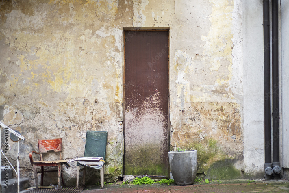 Fototapeta premium Pot for flowers, two chairs, peeling paint door near abandoned grunge cracked wall