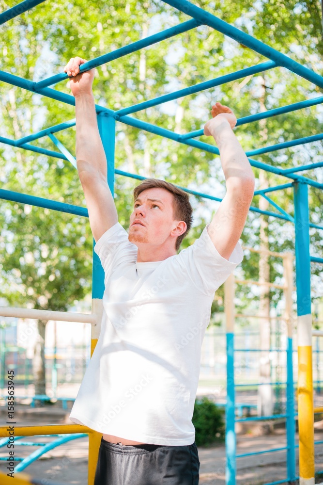 Obraz premium Strong young athlete. Strong young athlete is engaged in on an abandoned sports field. Athletic man. Exercises on uneven bars. Athlete Outdoors. Toned image.