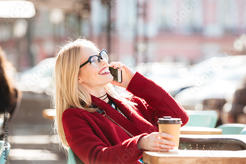 Happy young caucasian lady talking by phone drinking coffee.