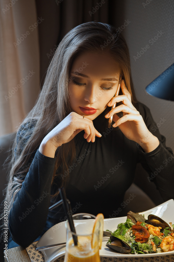 Young business woman sitting in the cafe and talking on the phone. Concept dinner. Orange juice and seafood salad. Indoor Young business woman sitting in the cafe and talking on the phone. Concept dinner. Orange juice and seafood salad. Indoor