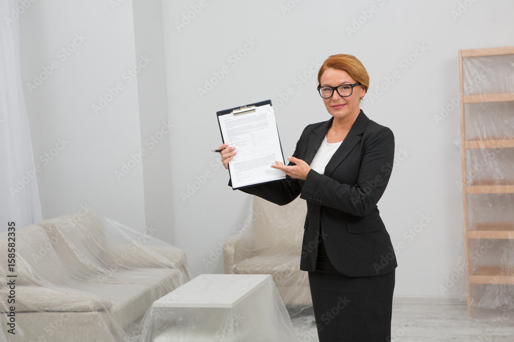 Agreemetn for apartment rent. Realtor agent standing in white room keeping an agreement for rent in her hands. Living room with furniture covered with foil. Agreemetn for apartment rent. Realtor agent standing in white room keeping an agreement for rent in her hands. Living room with furniture covered with foil.