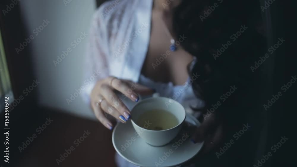 Morning of young beautiful bride. Close-up view of woman in lingerie, peignoir staying near window and drinking tea.