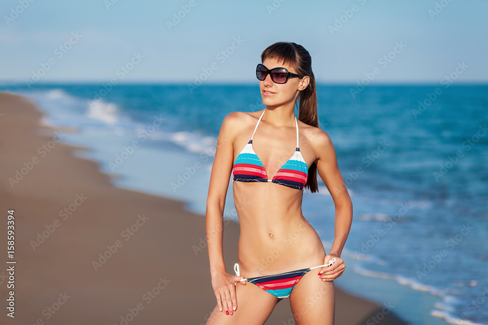Fashion outdoor portrait young sexy lady having fun and posing in striped bikini and sunglasses on sea background. Sunset light.