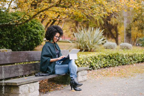 Stylish black female entrepreneur working with modern convertible laptop outdoor in autumn at city park. Young professional woman outside using tablet.