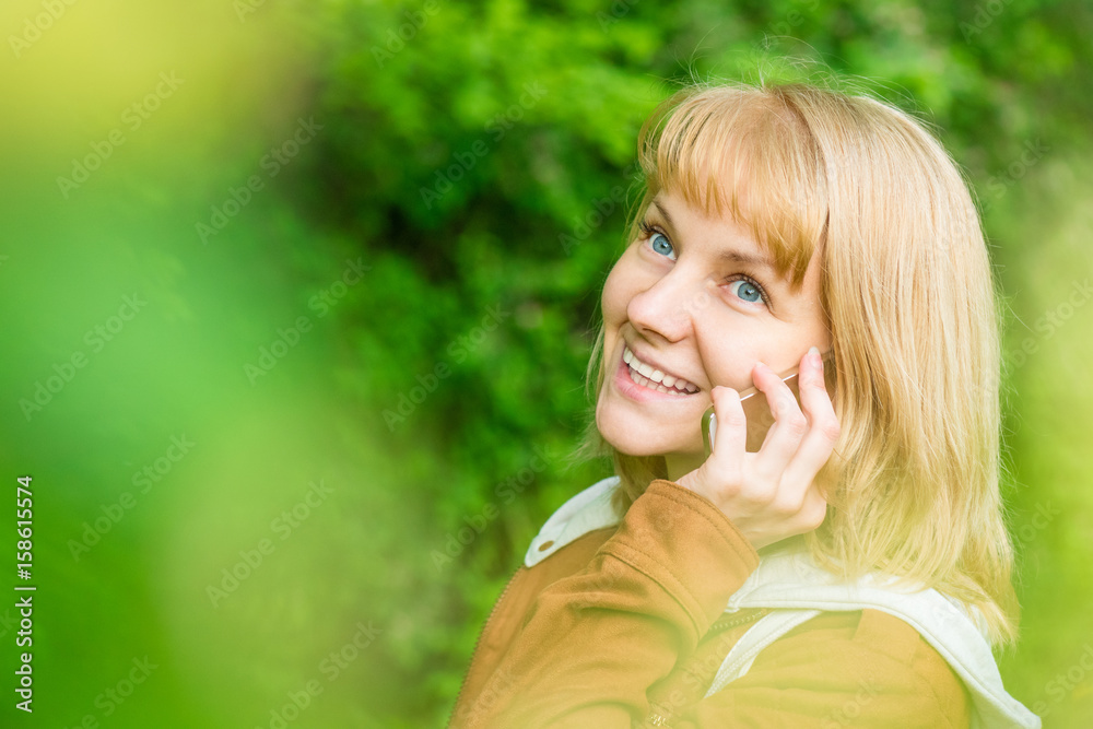 Naklejka premium Portrait of attractive smiling caucasian young woman with mobile, in spring park, looking away - close up. Beautiful girl talking on cell phone.