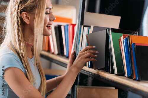 Smiling young woman choosing book on bookshelf in library