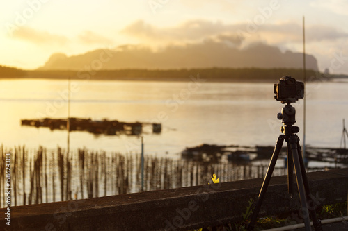 Camera set on a tripod aimed at a silhouette of a landscape at sunrise.