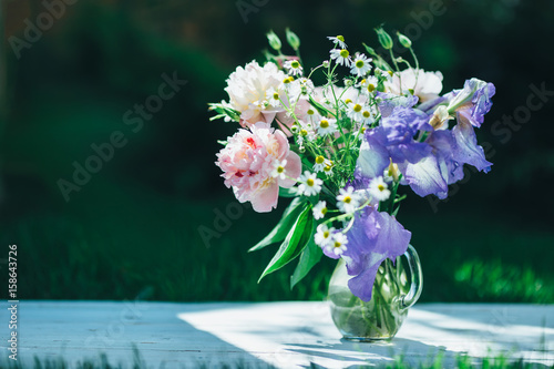 Bouquet of white peonies, chamomiles and iris flowers in glass vase. Summer background. Tinted photo
