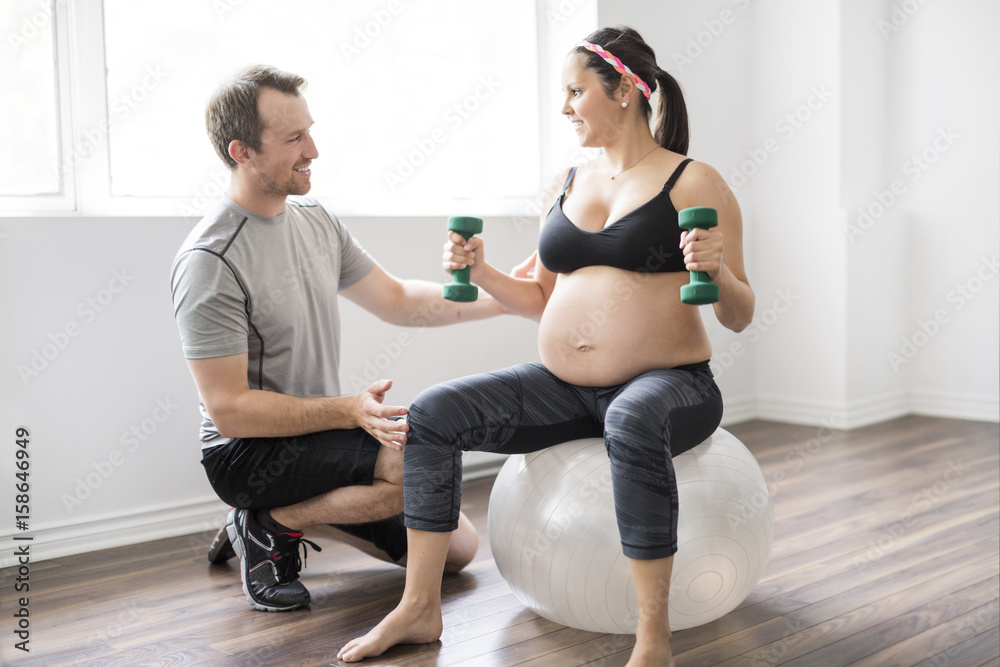 Obraz premium Pregnant woman working out with dumbbells with personal trainer at the gym