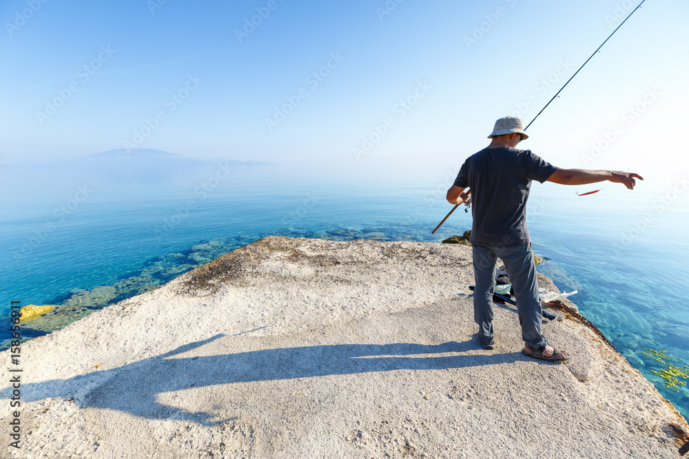 Side view of young man fisherman standing on pier with rod. Seashore of Ionian sea, Zante - Zakinthos island, Greece. Fishing background. Sunrise morning scenery.