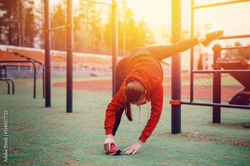 The girl on the sports ground stretches the muscles of her legs and back. The concept of a healthy lifestyle and exercises to strengthen the hands and back and joints. © Parilov