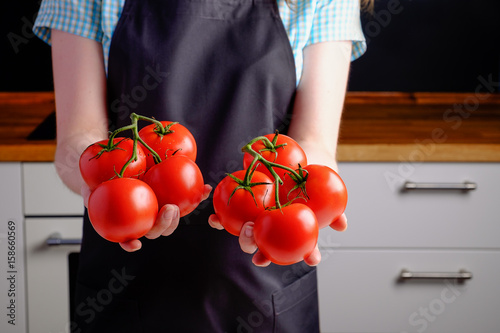 woman holding tomatoes in both hands at kitchen background. in black apron