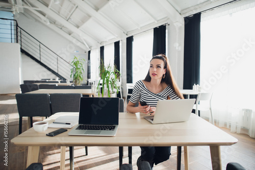 Adult woman at a modern office desk, working on laptop and phone, looking away, thinking about a post, full-time blogger, seeking for inspiration. photo
