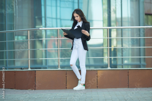 Beautiful young girl in a suit with a black folder in hands on the background of a glass building with a pensive expression