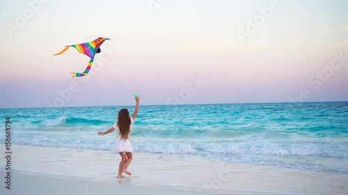 Little girl with flying kite on tropical beach. Kid play on ocean shore. Child with beach toys in slow motion