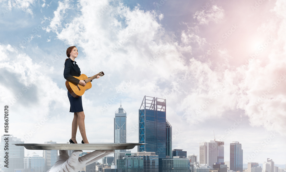 Attractive businesswoman on metal tray playing acoustic guitar against cityscape background Attractive businesswoman on metal tray playing acoustic guitar against cityscape background