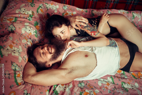 Loving couple lying in bed together. Handsome young hipster with beard and attractive woman resting at home while kissing and cuddling.