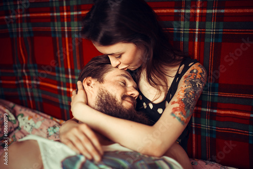 Loving couple lying in bed together. Handsome young hipster with beard and attractive woman resting at home while kissing and cuddling.