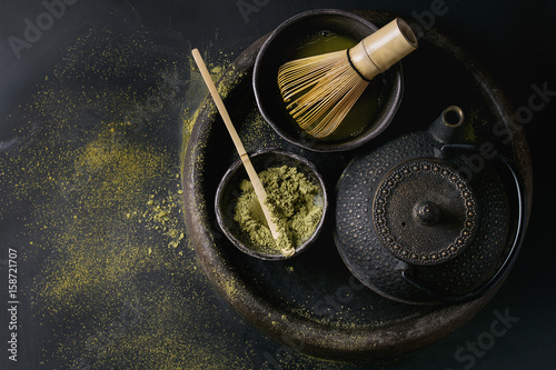 Green tea matcha powder and hot drink in black bowls standing with iron teapot, bamboo traditional tools spoon and whisk in terracotta tray over dark metal background. Top view with space