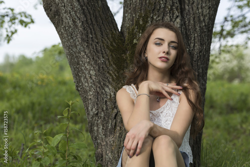 A girl with long hair sits, leaning her back on the trunk of a tree