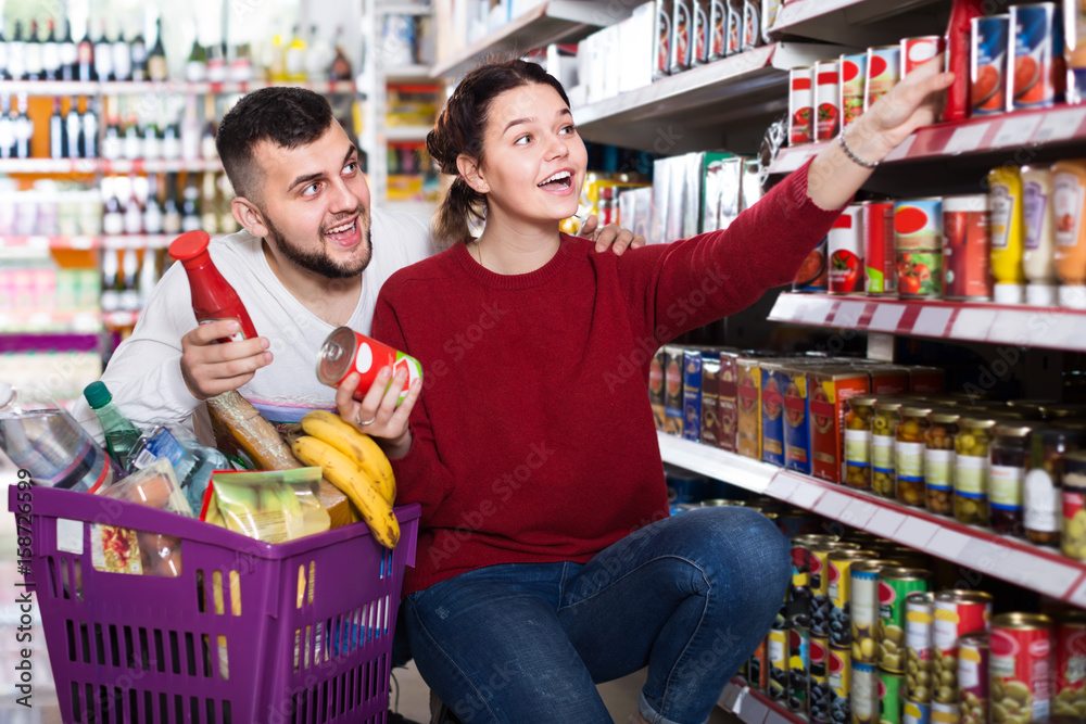 smiling young couple choosing purchasing canned food for week at supermarket